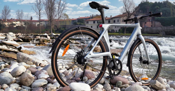 Fiido Air ultralichte elektrische fiets van koolstofvezel, opgesteld op een rotsachtige rivieroever naast stromend water.