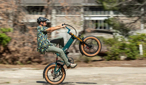 Een fietser maakt een wheelie op een Fiido M1 Pro fatbike en laat op een stedelijke weg het compacte vouwframe, de brede banden en de voorvering zien.