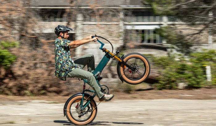 Een fietser maakt een wheelie op een Fiido M1 Pro fatbike en laat op een stedelijke weg het compacte vouwframe, de brede banden en de voorvering zien.