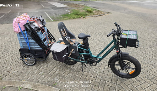 Een Fiido T2 elektrische bakfiets is volledig beladen met een kinderzitje, zijtassen, een voorste laadbak en een zwaar beladen aanhanger.